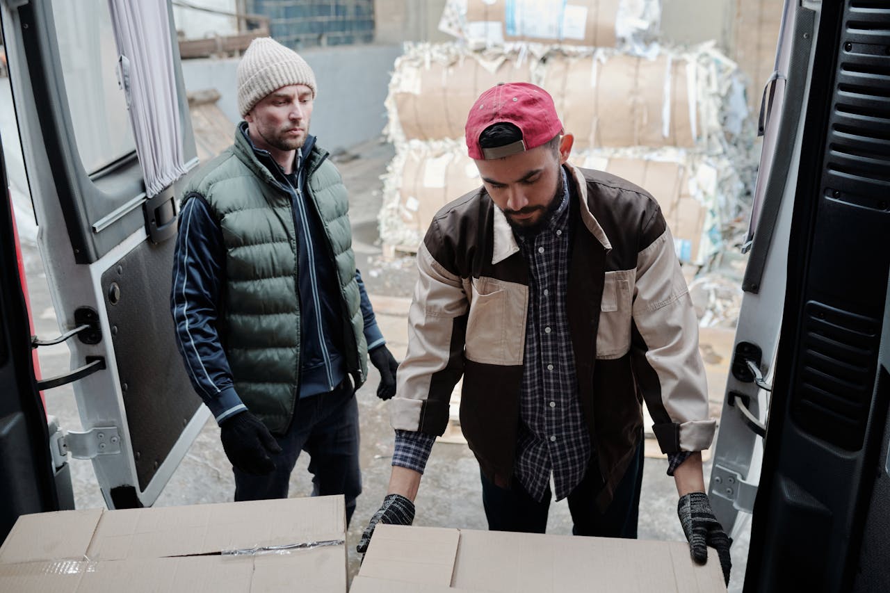 Two men loading cardboard boxes into a delivery van at an urban site.