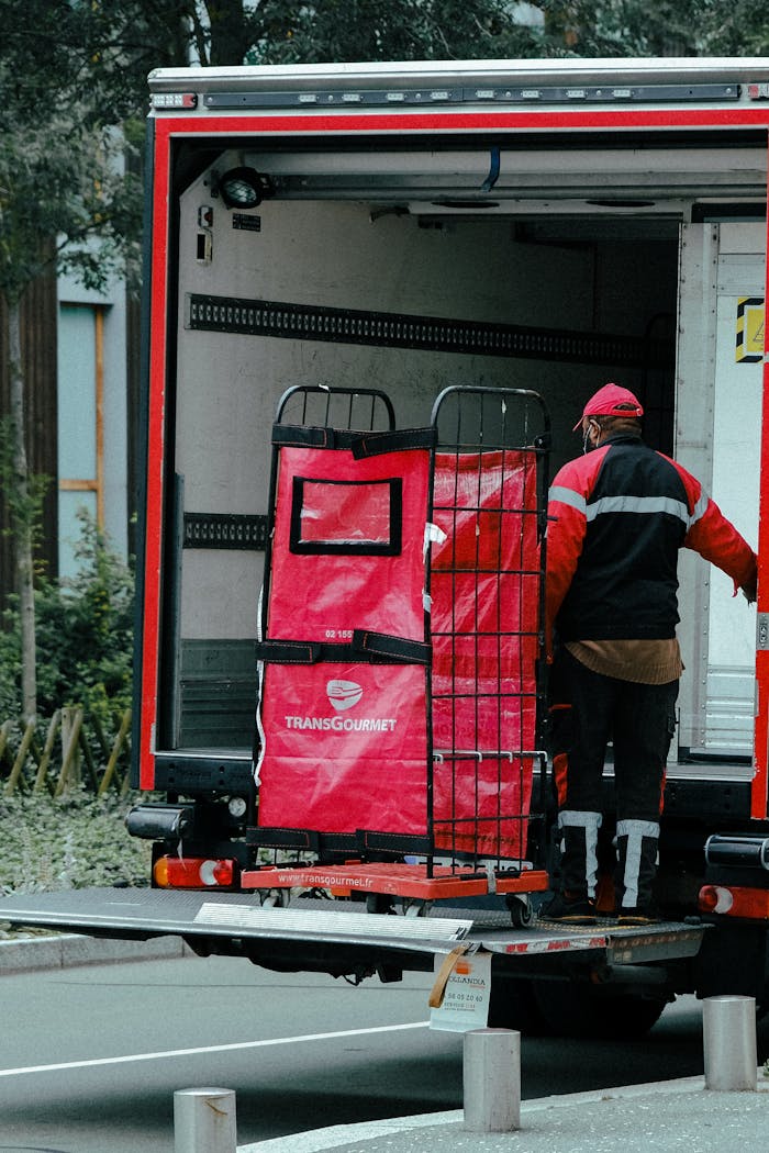 services-04 A delivery man is unloading a truck filled with supplies on an urban street, wearing a red and black uniform.